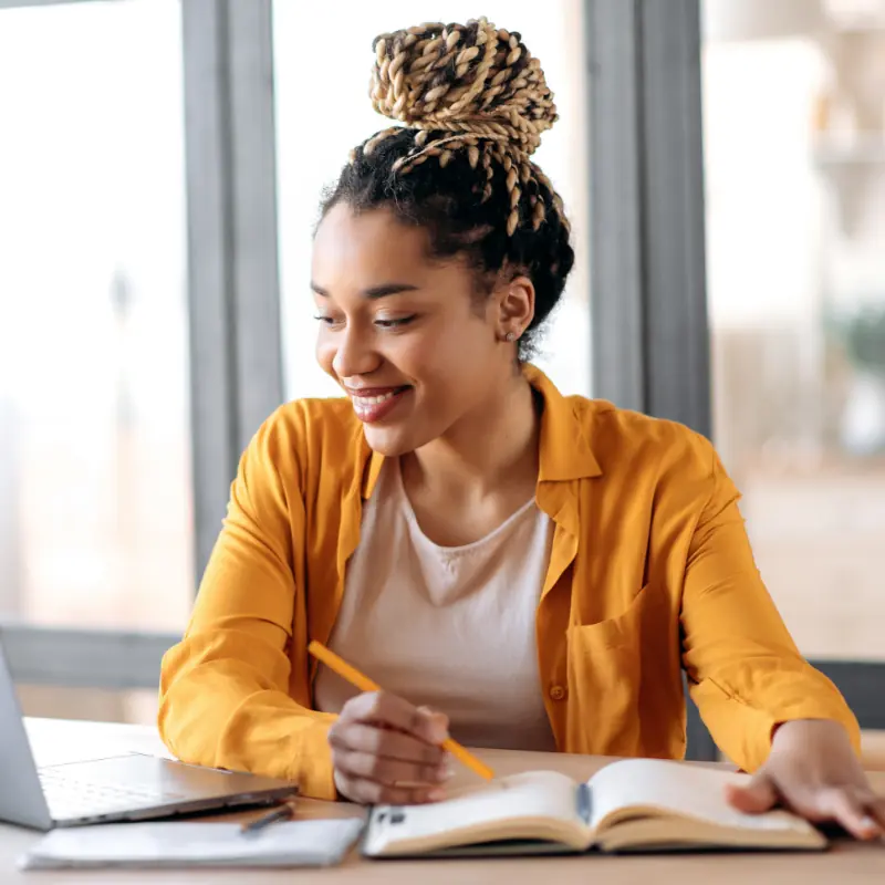 Online education, e-learning. Happy african american young woman in stylish casual clothes, studying remotely, using a laptop, listening to online lecture, taking notes while sitting at home, smiles