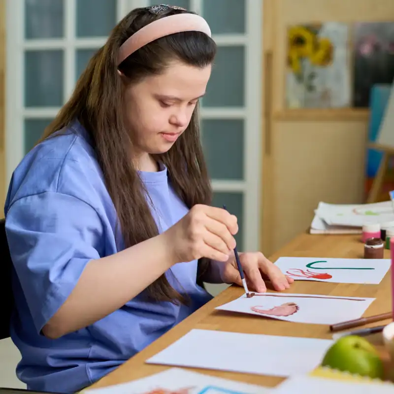 Girl with mental disability painting with watercolors and paintbrush on paper while sitting by desk in front of laptop in living room